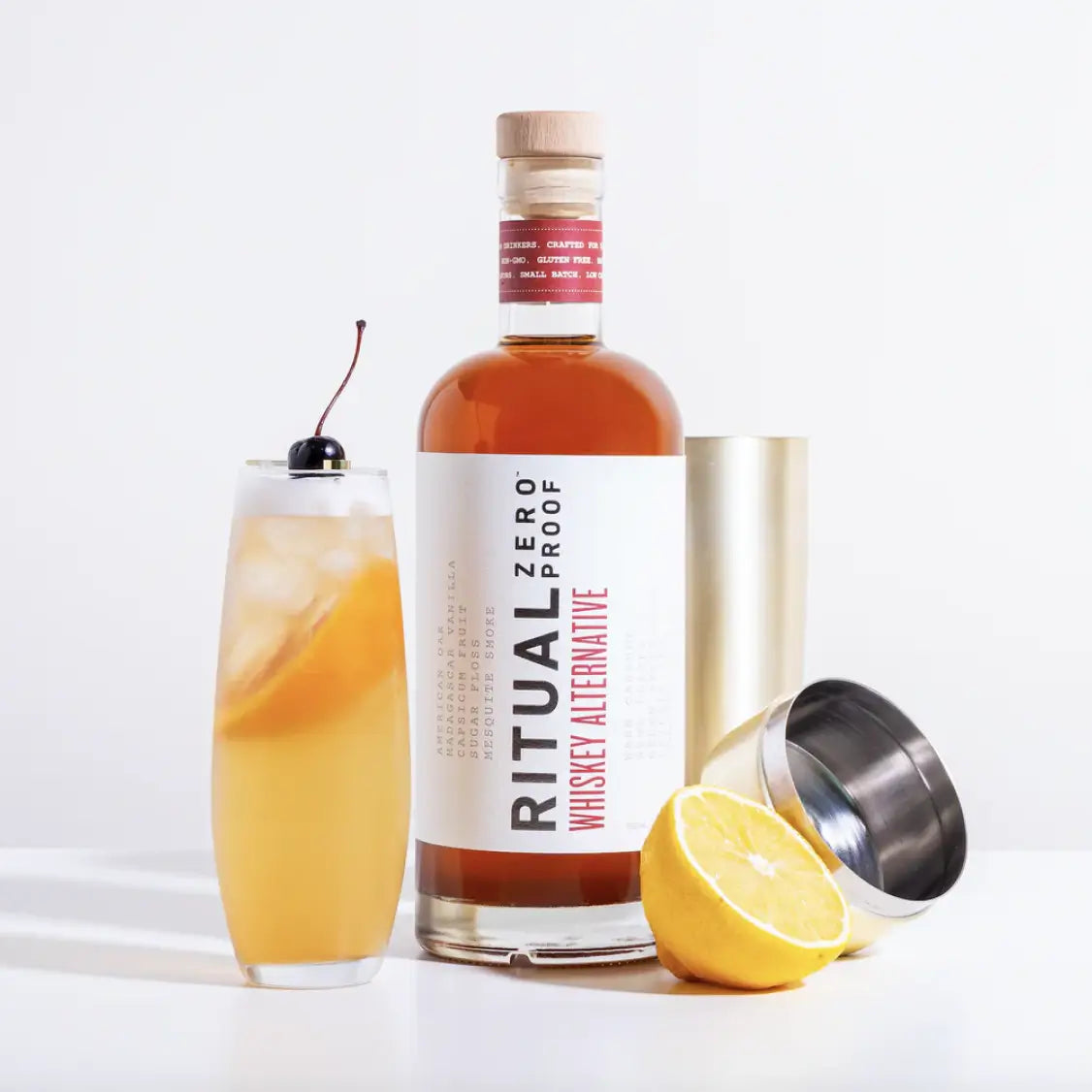 Bottle of Ritual Zero Proof Whiskey Alternative with a cocktail, lemon half, cherry garnish, and cocktail shaker.