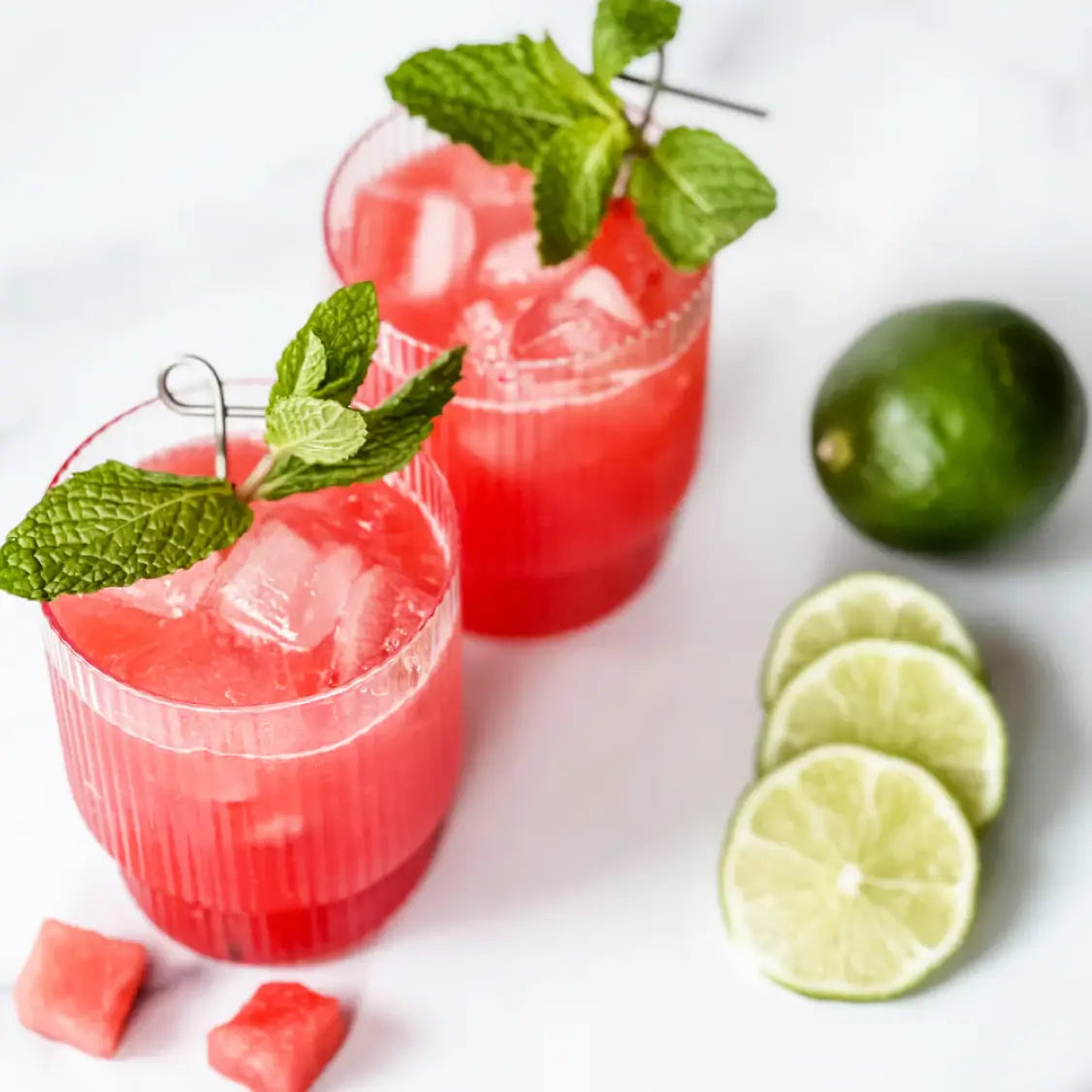 Two non-alcoholic watermelon smash cocktails with ice and mint garnish, served alongside lime slices and watermelon cubes.